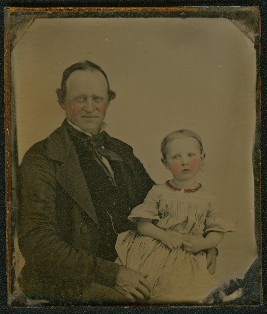 A vintage portrait of an older man and a young girl, both with rosy cheeks. The man wears a dark suit, and the girl wears a light-colored dress with a red necklace. She sits on his lap, both gazing towards the camera. The photograph has an antique feel.