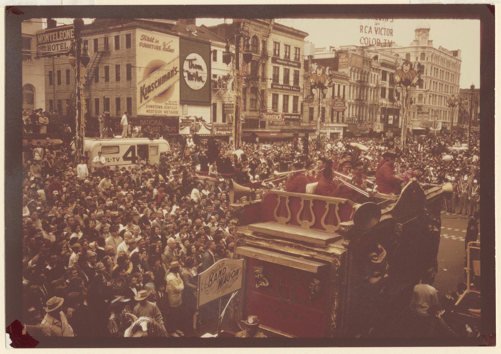 A vibrant parade scene with a large crowd gathered on the street. A float with musicians playing instruments is in the foreground. Historic buildings and various signs line the background, including one for a hotel.