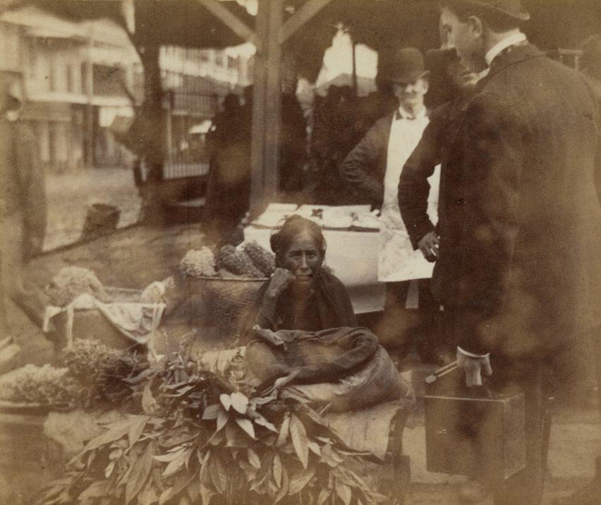A sepia-toned historical photo of a street market scene. A woman sits behind a display of produce, including leafy greens. She is surrounded by three men in suits and hats, one carrying a briefcase. The background shows a table and buildings.