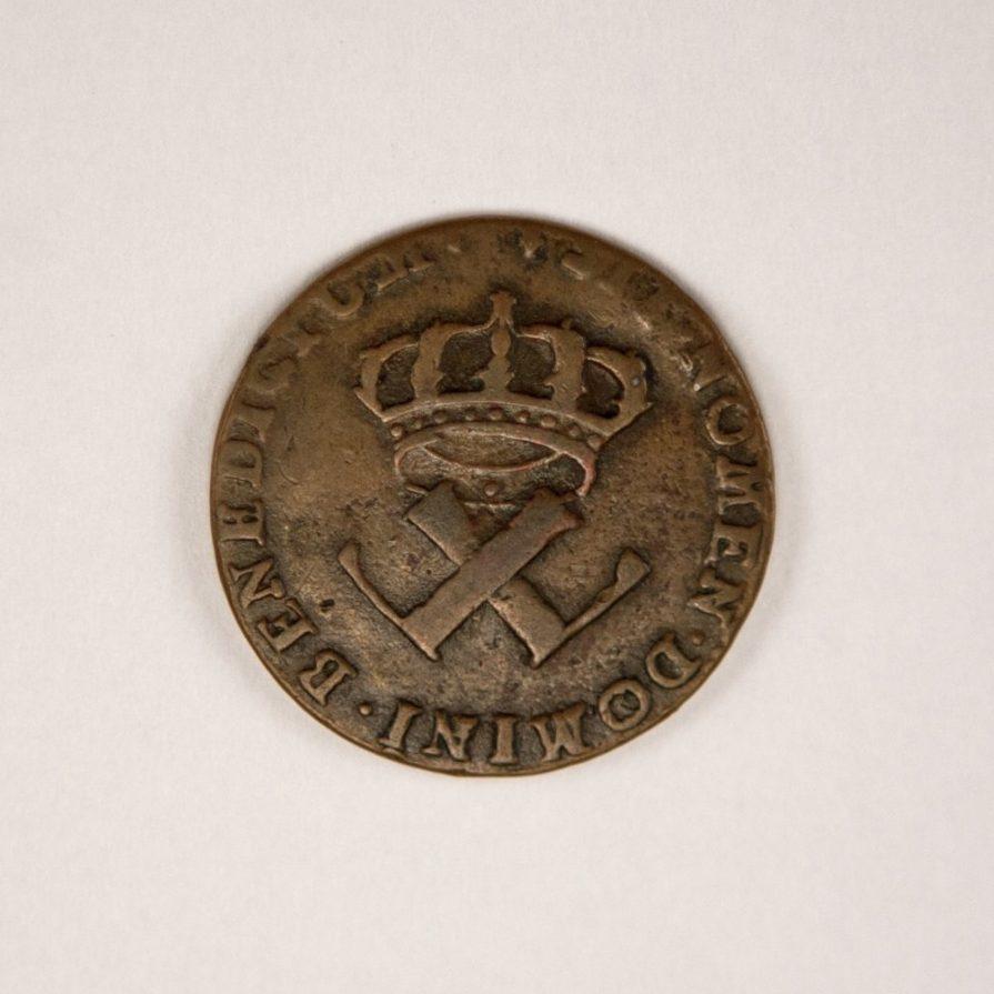 An old coin with a crowned emblem featuring intertwined tools, possibly axes, and the inscription BENEDICTUM MOMENTUM DOMINI around the edge. The coin has a worn and tarnished appearance.