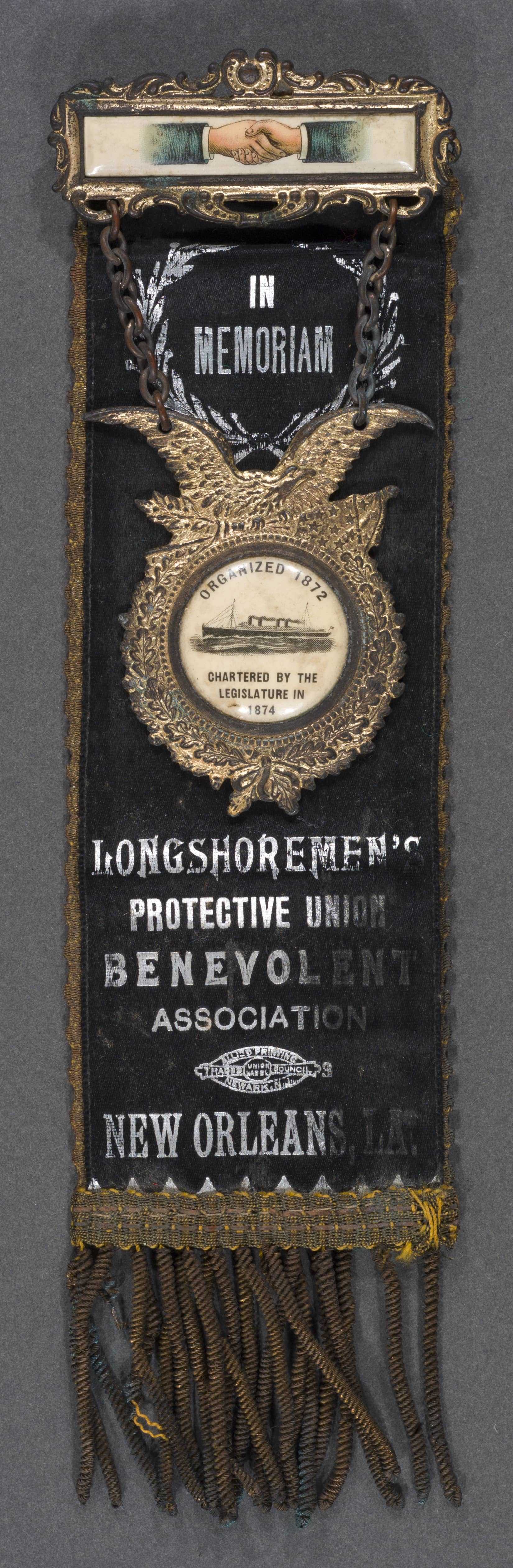 A vintage badge with In Memoriam at the top. Below, theres an emblem featuring an eagle, with text indicating affiliation to the Longshoremens Benevolent Association, New Orleans, LA. Tassels hang from the bottom.