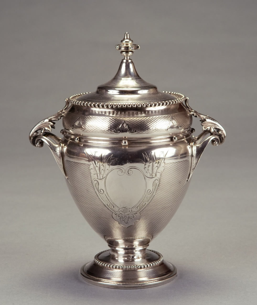 A silver urn with a decorative lid, featuring intricate engravings and two ornate handles. Its surface has a textured pattern and a classic, elegant design, resting on a round base against a plain background.