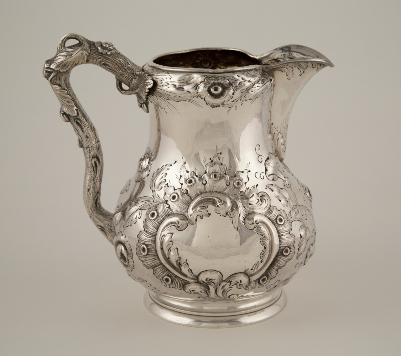 Antique silver pitcher with ornate floral and scroll designs on its body and handle, set against a plain background. The intricate detailing highlights craftsmanship and elegance.