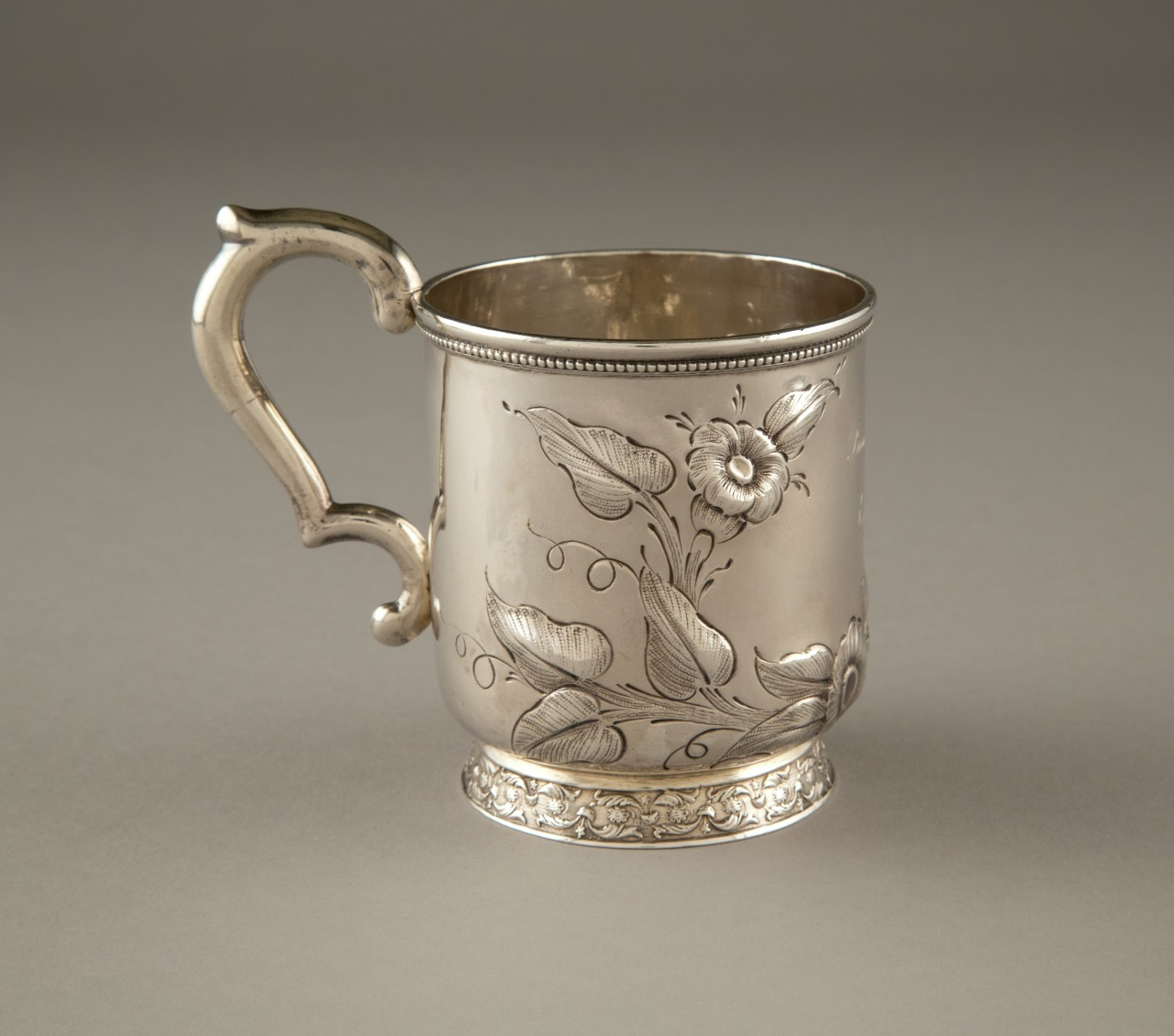 A silver mug with an intricately engraved floral pattern and a decorative handle, set against a plain background.