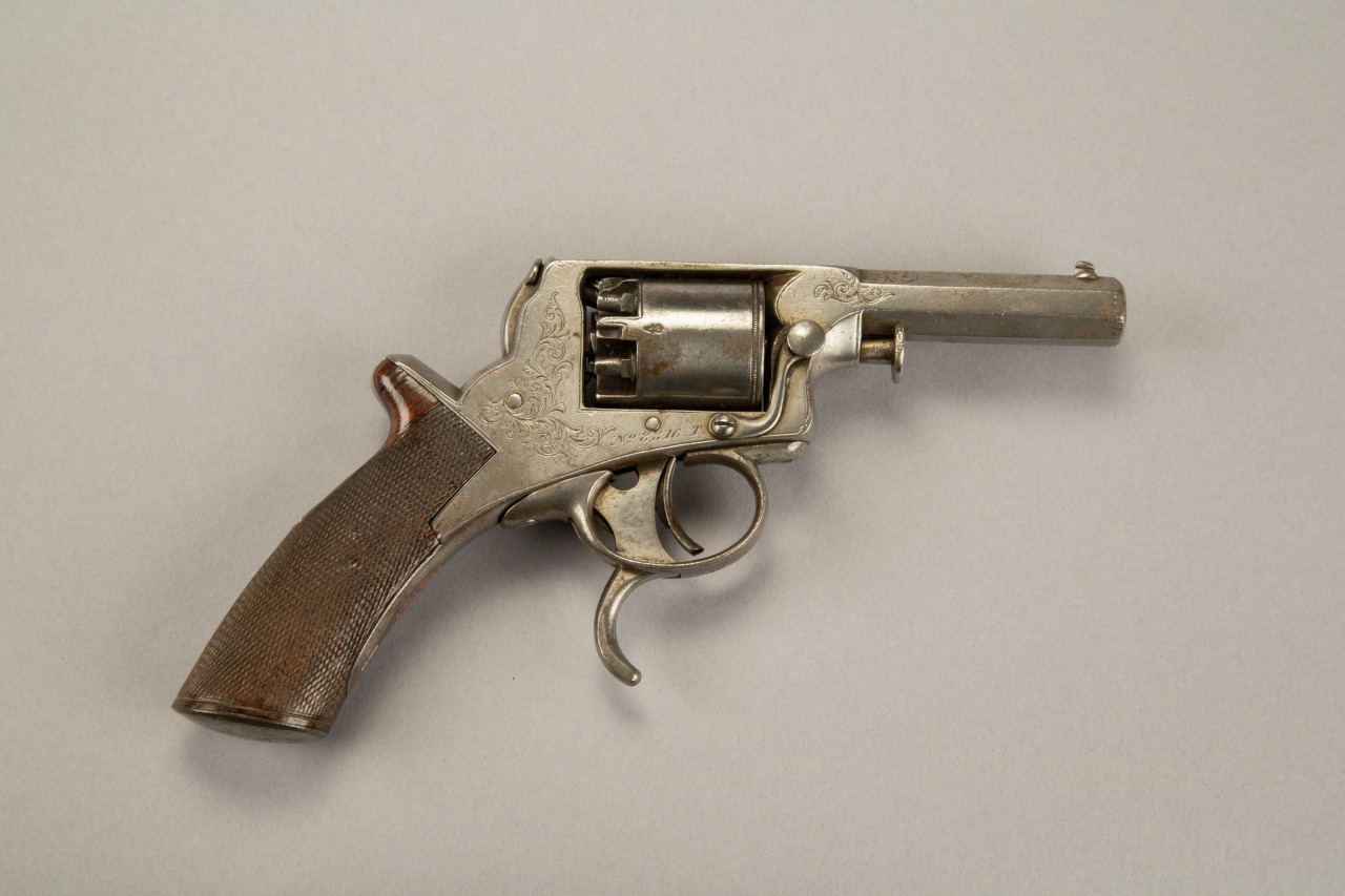 Antique revolver with a wooden grip and metal frame, featuring a closed cylinder and a short barrel. The gun is placed on a plain gray background.