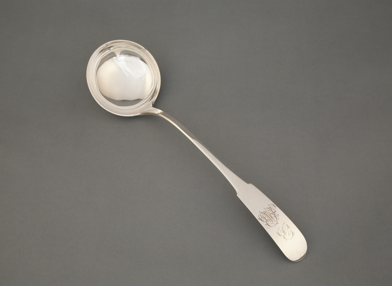 A silver ladle with an engraved handle is placed on a gray background. The ladle has a round, deep bowl and a long handle with ornate detailing near the top.