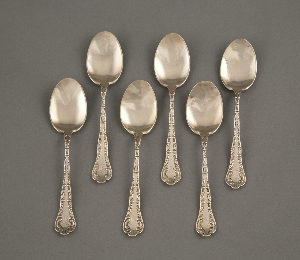 Six ornate silver spoons with intricate designs on the handles are arranged in two rows on a gray background.