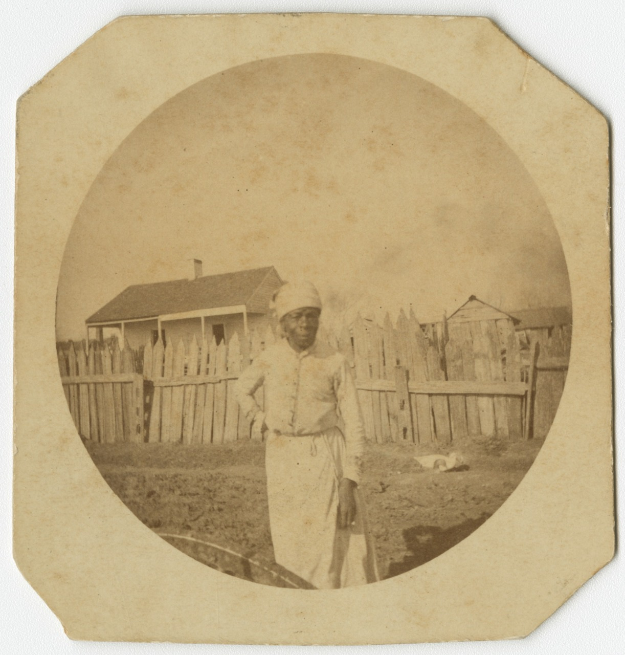A woman in 19th-century clothing stands outside in front of a wooden fence and small houses. She wears a headscarf and apron, with her hand resting on her hip. The photograph has a sepia tone, typical of the era.