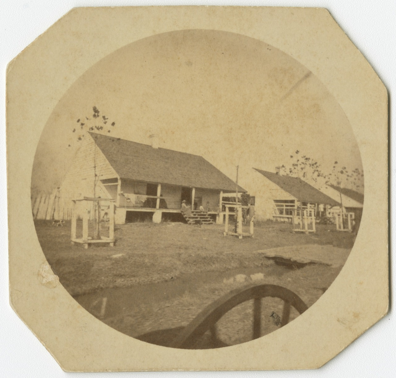 Sepia-toned photograph of a rural scene with two wooden cabins in the background. The cabins have porches and sloped roofs. In the foreground, part of a wooden wagon wheel is visible. Sparse trees are scattered around the area.