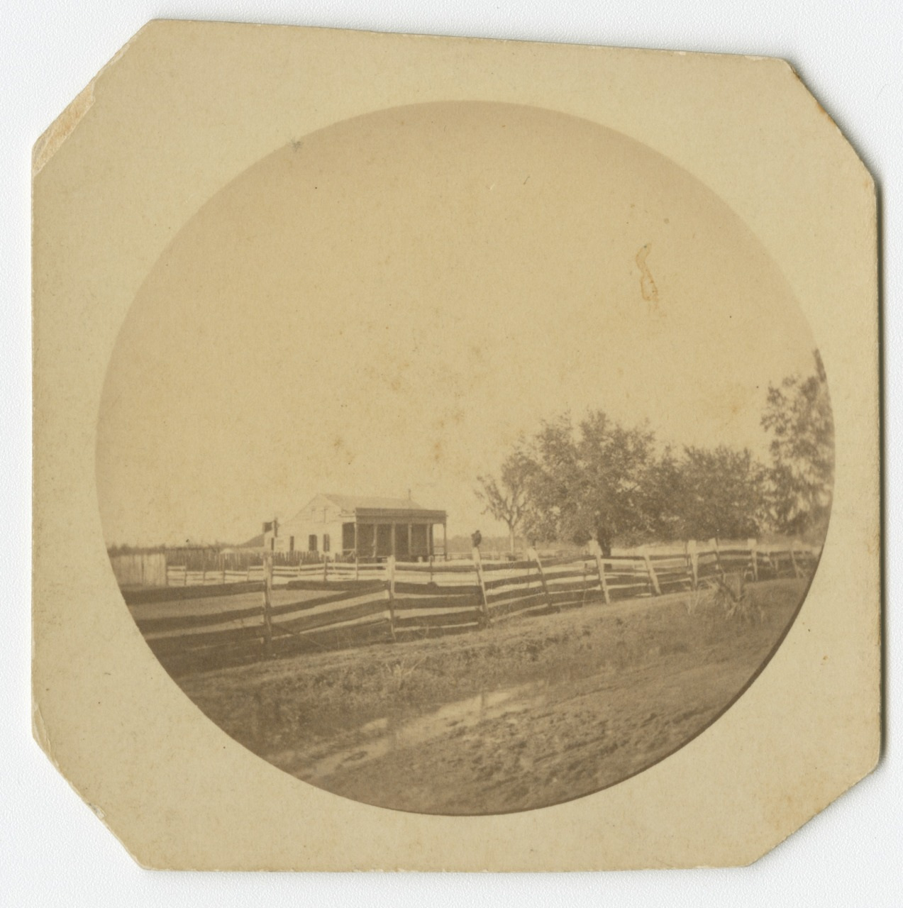 Sepia-toned vintage photograph showing a small house on a grassy field, with a wooden fence stretching across the foreground and several trees nearby under a clear sky.
