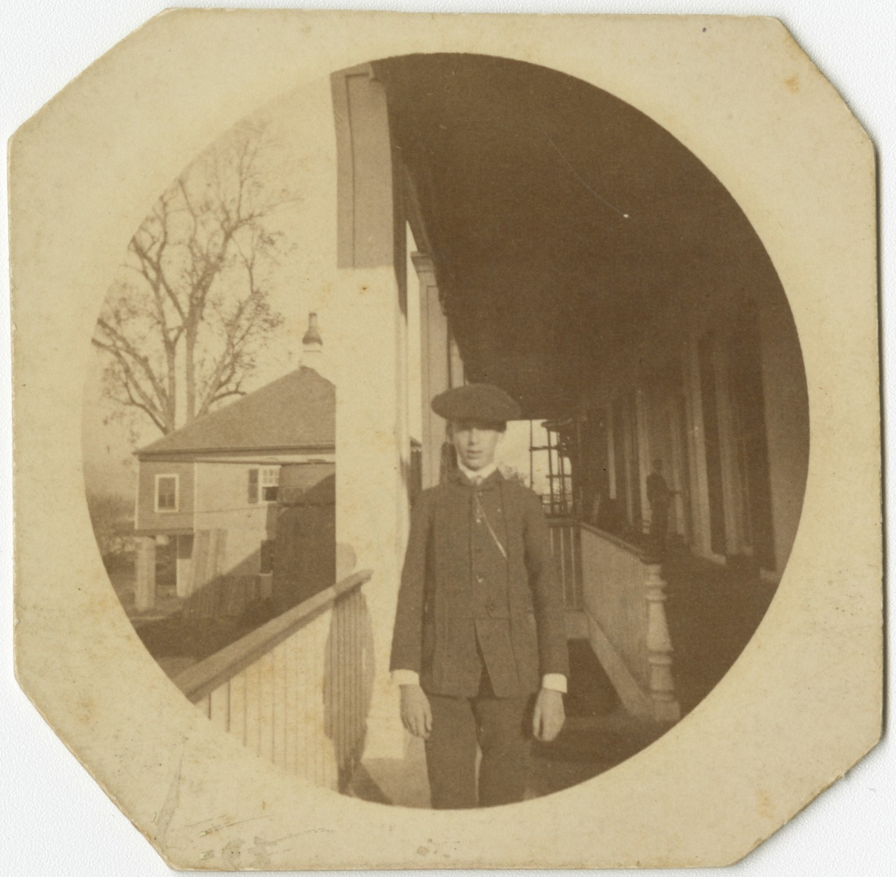 Vintage sepia photograph of a young person wearing a suit and cap, standing on a porch. A large tree and a house are visible in the background. The photo has rounded edges and an old-fashioned border.