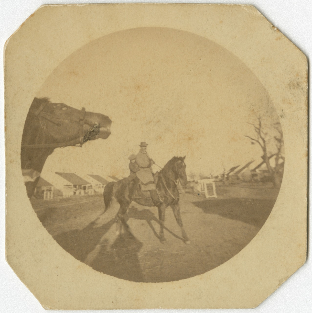 Sepia-toned photograph of a person riding a horse, with another horses head prominently in the foreground. The scene is set on a dirt road, surrounded by tents and sparse trees. The overall image has a vintage and historical feel.