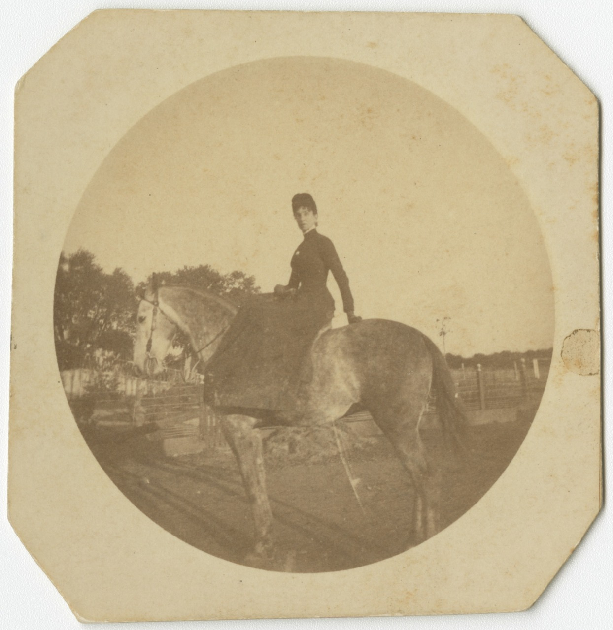 A sepia-toned vintage photo of a person on horseback. The rider is dressed in traditional attire and poses side-saddle. The background features a rustic outdoor setting with fences and trees. The image has worn edges, indicating its age.