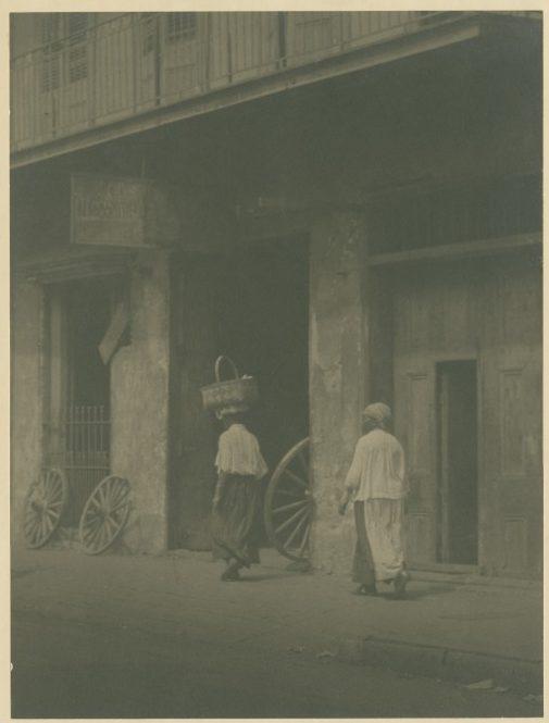Two people in traditional clothing walk down a dimly lit street beside an old building. One carries a basket on their head. Wooden wheels lean against the wall, and a balcony is visible above. The scene is captured in a vintage, sepia-toned style.