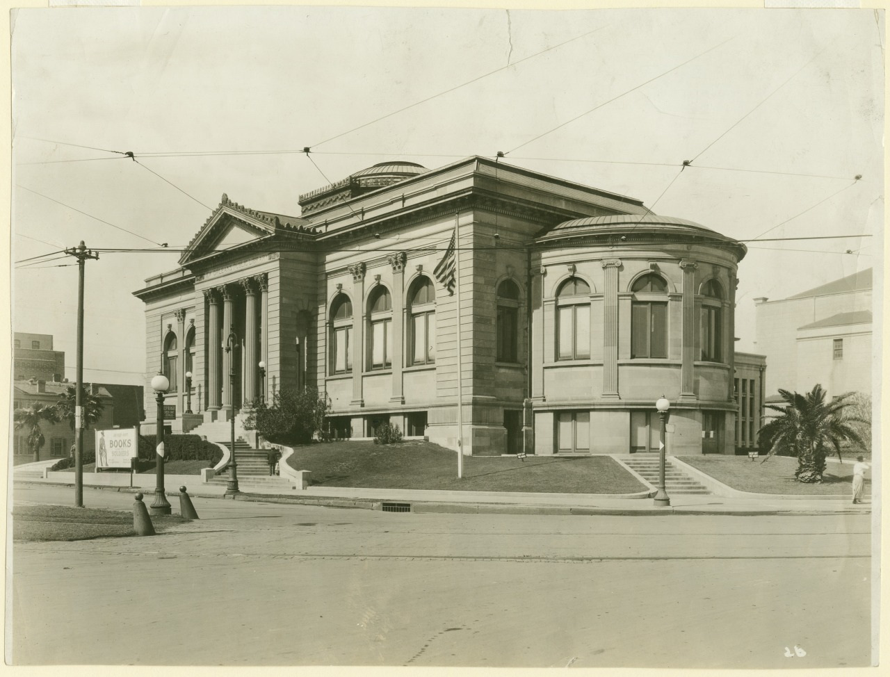 A vintage black and white photograph of a large, historic building with classical architecture, featuring tall columns, arched windows, and an ornate pediment. It is set on a grassy area with steps leading up to the entrance.