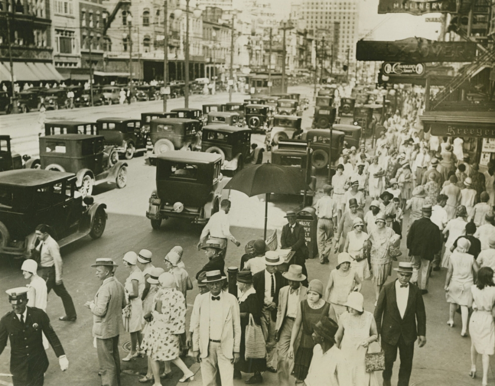 A bustling city street scene from the early 20th century, showing people in period attire and hats walking on a crowded sidewalk. Numerous vintage cars are on the road, with shops and buildings lining the street.