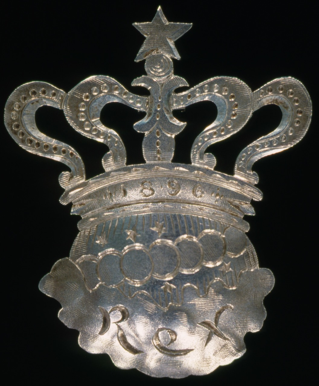 A silver emblem of an ornate crown with a star on top and the text Rex at the bottom. The crown features intricate details, including scrolling elements and a series of circles in the center, marked with the year 1896.