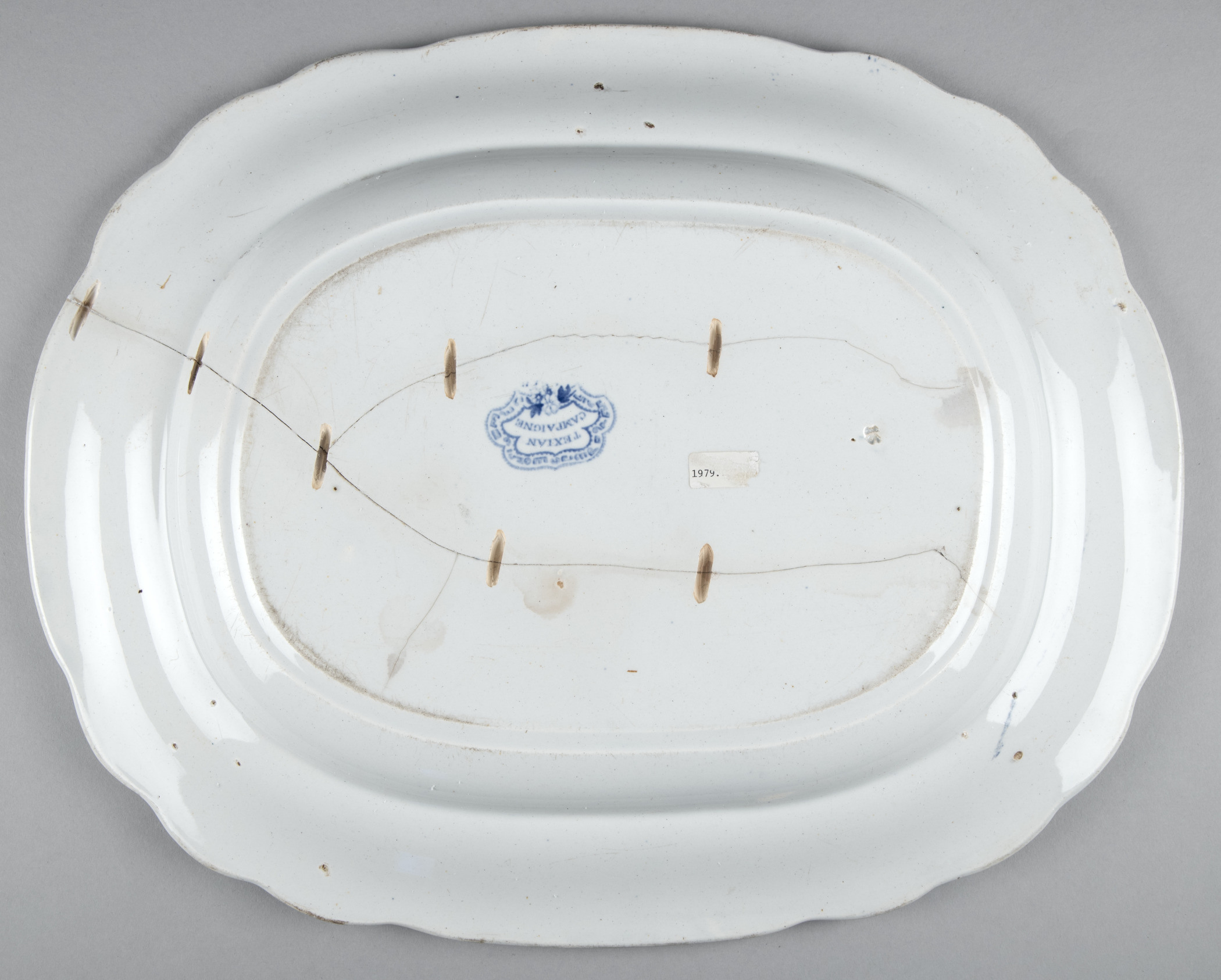 The image shows the back of an oval white ceramic platter with a scalloped edge. It features a blue emblem with the word Windsor and has several visible repair wires attached. The platter has some stains and a small label.