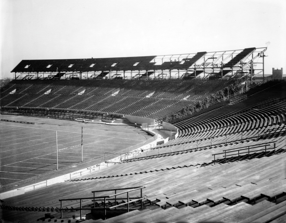 Black and white photo of a partially constructed sports stadium. Empty stands curve around the field, with visible framework for the roof. The field has markings for a football game, and no people are present.