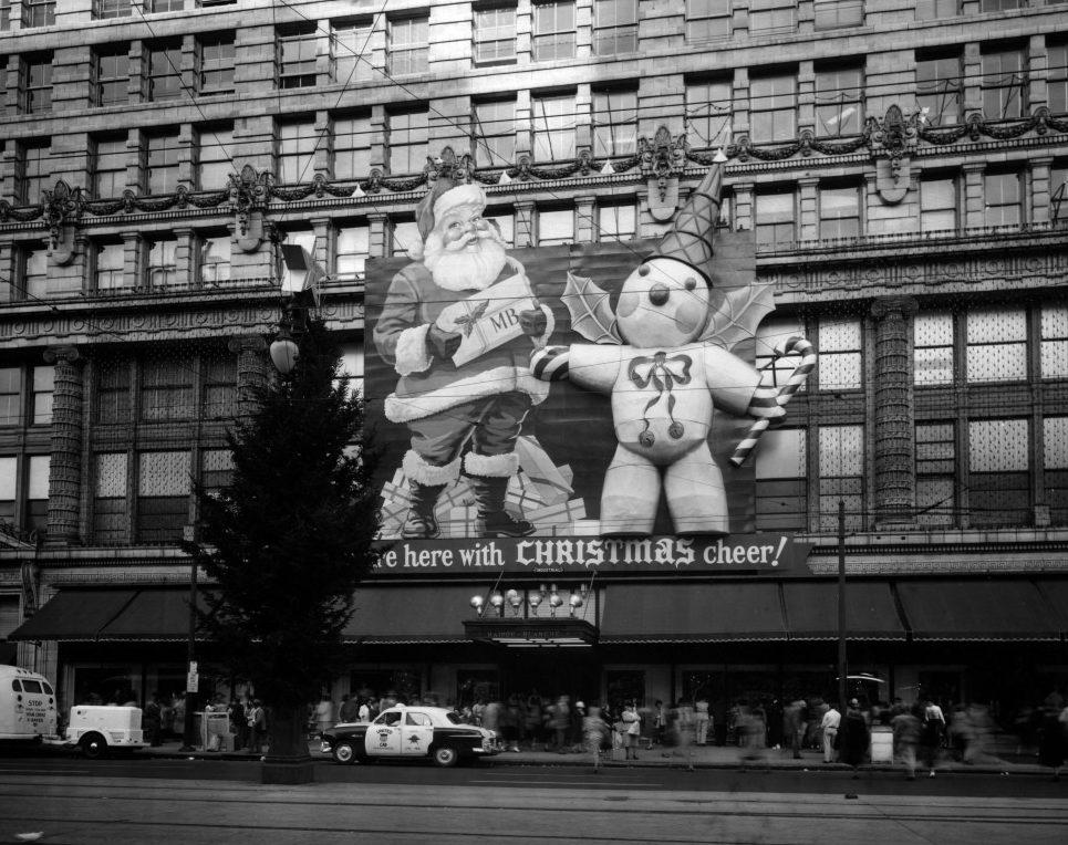 A black and white photo of a department store decorated with large inflatable Santa Claus and snowman figures. A Christmas tree stands in front, with people and cars visible on the street below.