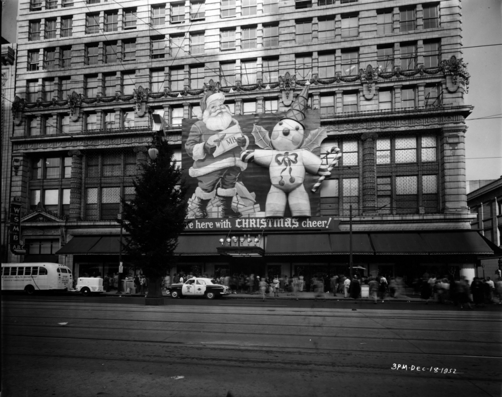 Black and white photo of a large department store decorated for Christmas with giant Santa and snowman cutouts. A tree stands outside, and people walk by. Buses and cars are on the street.