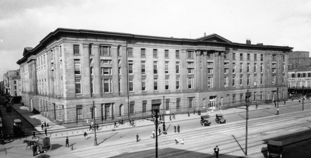 A historic black-and-white photo of a large, multi-story stone building with numerous windows. People and early 20th-century cars are visible on the streets surrounding the building. Power lines cross the scene.