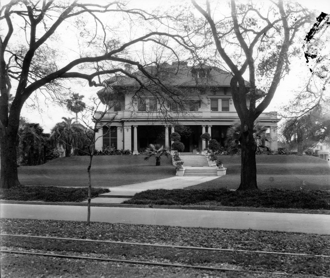 A historic, two-story mansion with a wraparound porch is surrounded by large trees. A manicured lawn leads to the front steps and pathway. The street in the foreground has tram tracks. The photo is in black and white, giving it a vintage feel.