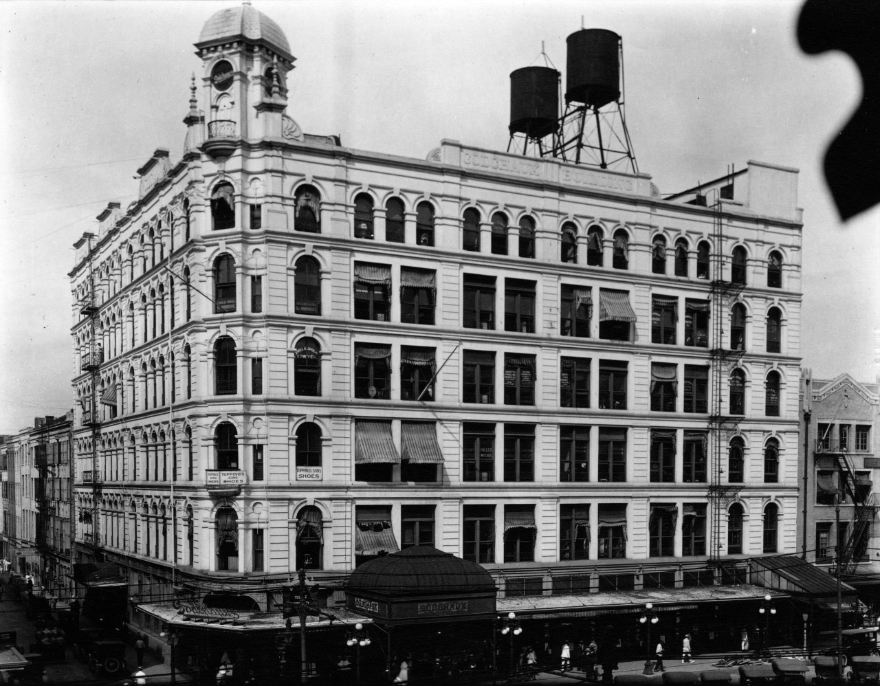 A historic black-and-white photo of a five-story building with ornate architecture and two water towers on the roof. Street-level shops and awnings are visible, and people are walking along the bustling sidewalk below.
