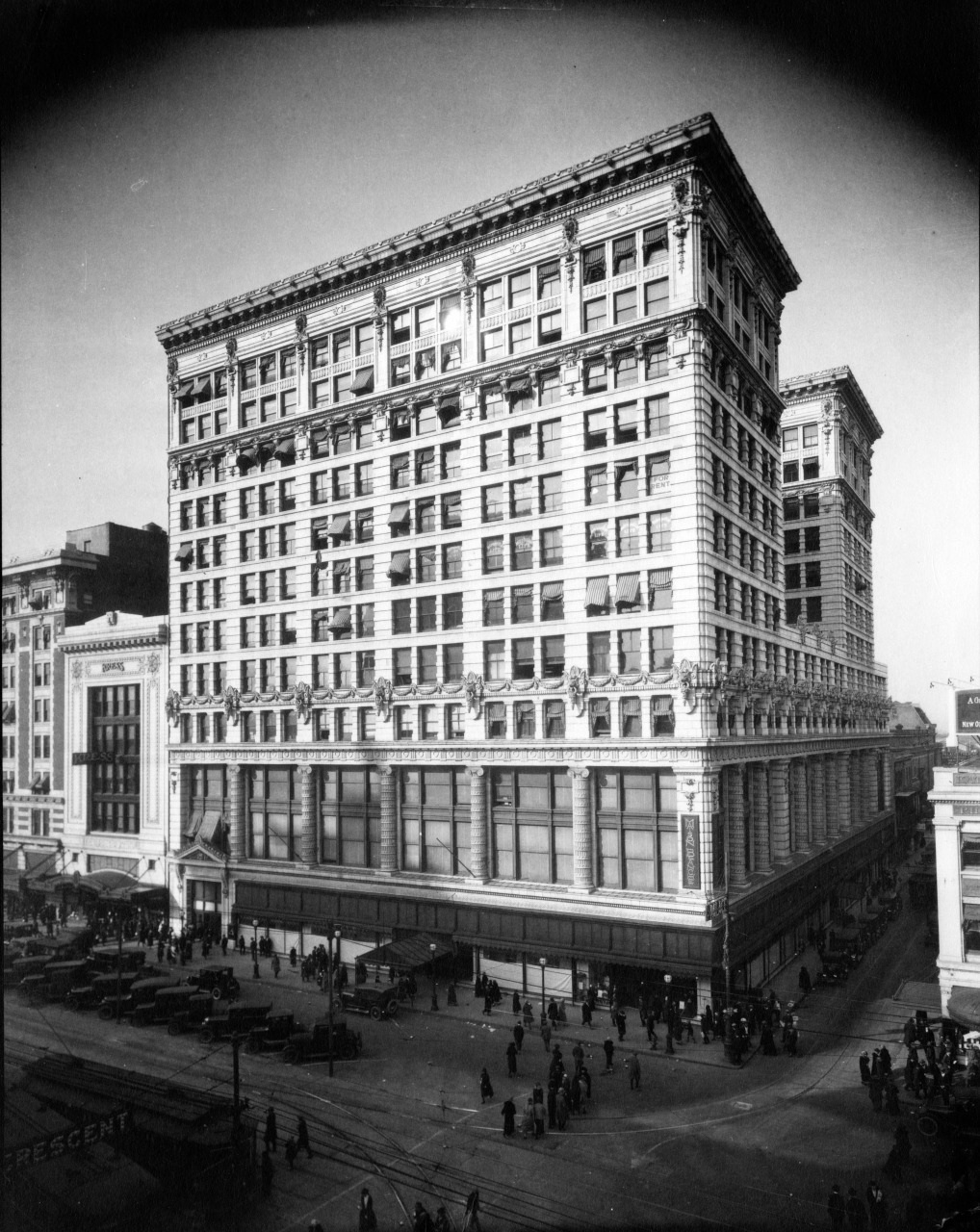 A black and white photograph of a bustling, early 20th-century city street featuring a large, ornate building with multiple stories. The street is filled with people and horse-drawn carriages.
