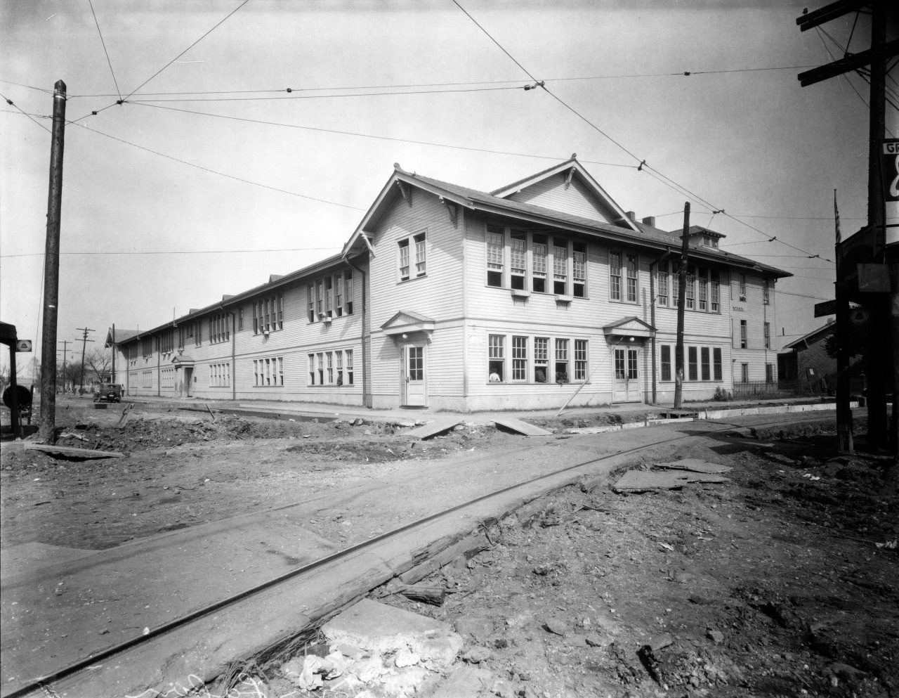 A large, two-story wooden building with numerous windows sits on a dirt-covered corner, surrounded by utility poles and wires. The scene is industrial and devoid of vegetation. The sky is overcast.