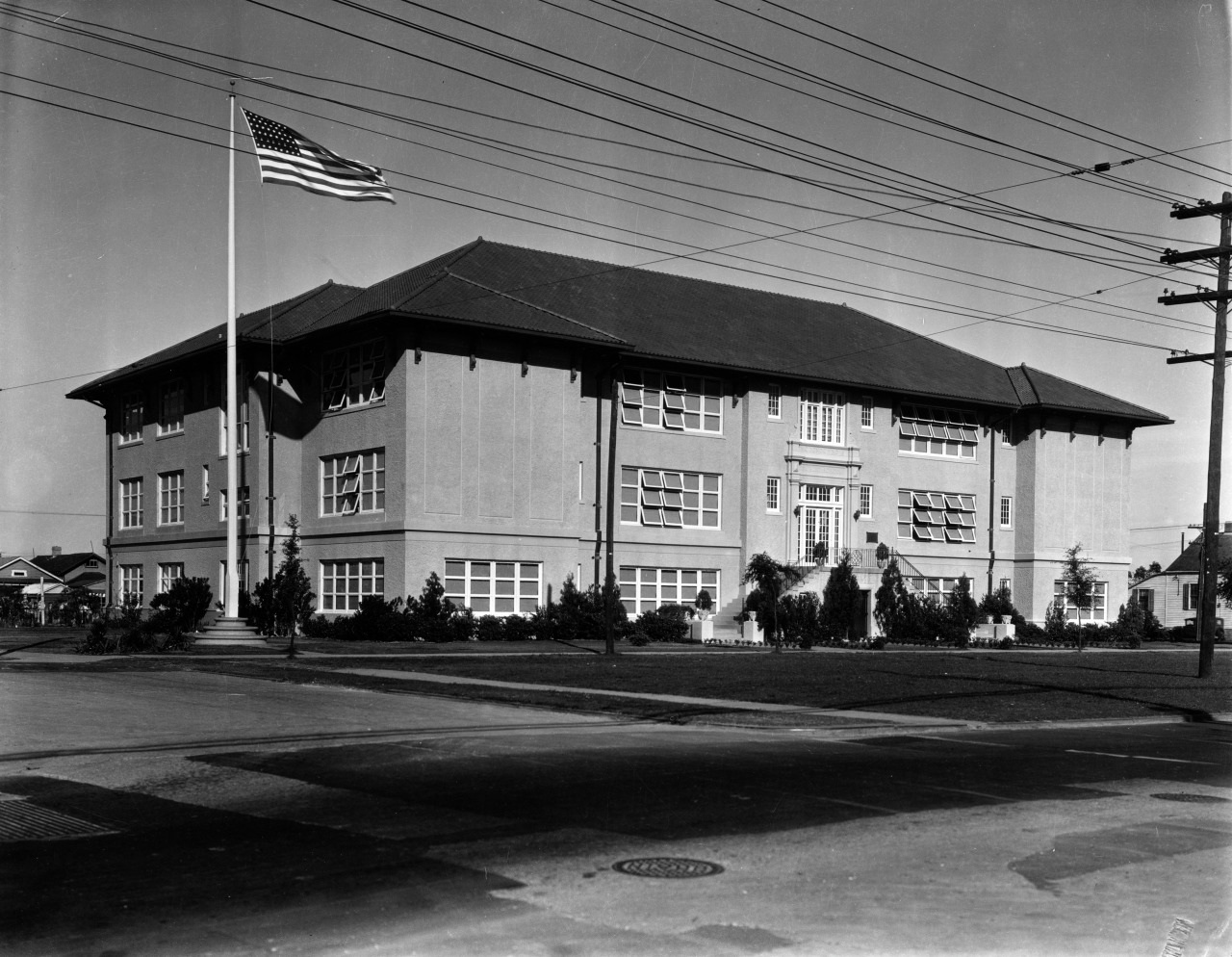 A large, two-story brick building with multiple windows sits at a corner. An American flag waves on a pole in the foreground. Power lines run overhead, and shrubs line the buildings perimeter. The sky is clear.