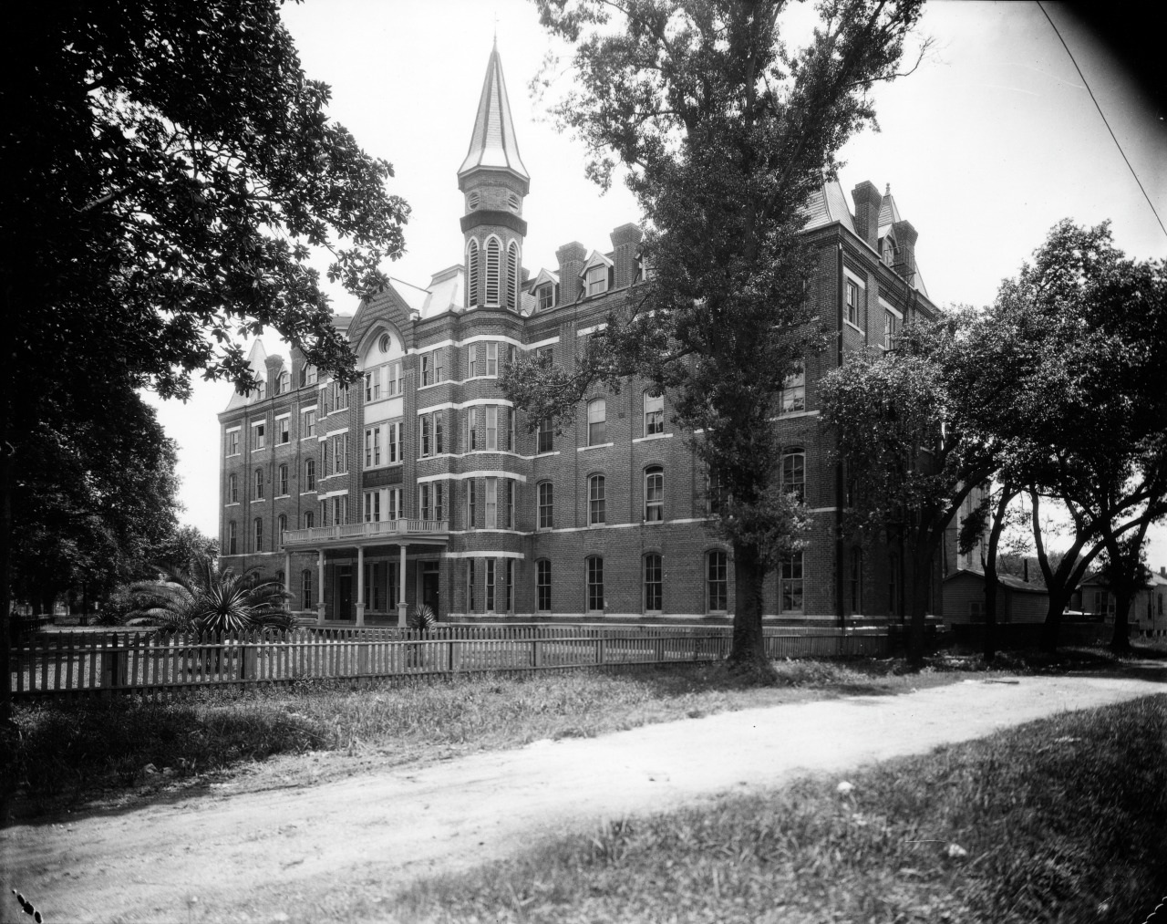 A historic, multi-story brick building with a tower and gabled roof stands behind a picket fence. Tall trees surround the structure, and a dirt path leads past it, evoking a vintage and timeless feel.
