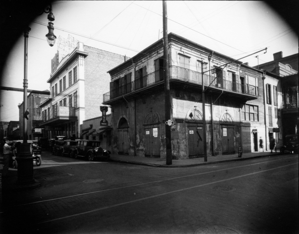 Black and white photo of a historic street corner in New Orleans, featuring vintage cars parked along the curb and a two-story building with a balcony. Signs and power lines are visible against a clear sky.