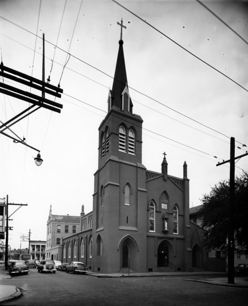 A historic church with a tall steeple and cross, situated at a street corner. The architecture features arched windows and doors. Several cars are parked nearby, and power lines crisscross overhead against an overcast sky.
