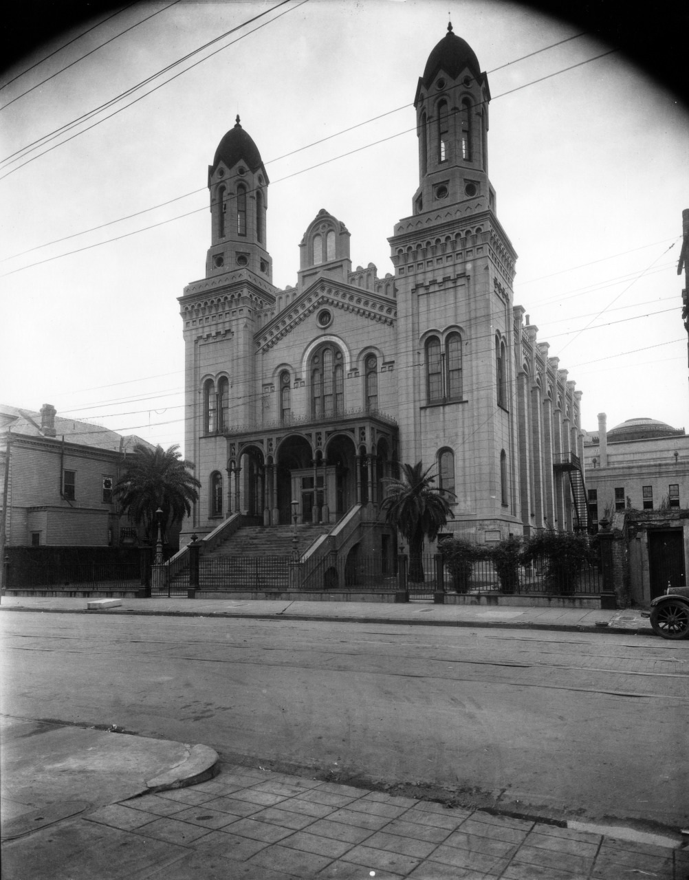 A historic cathedral with twin towers and ornate details stands behind a gated entrance. Palm trees flank the front steps, and a car is partially visible on the right. The scene captures early 20th-century architecture.