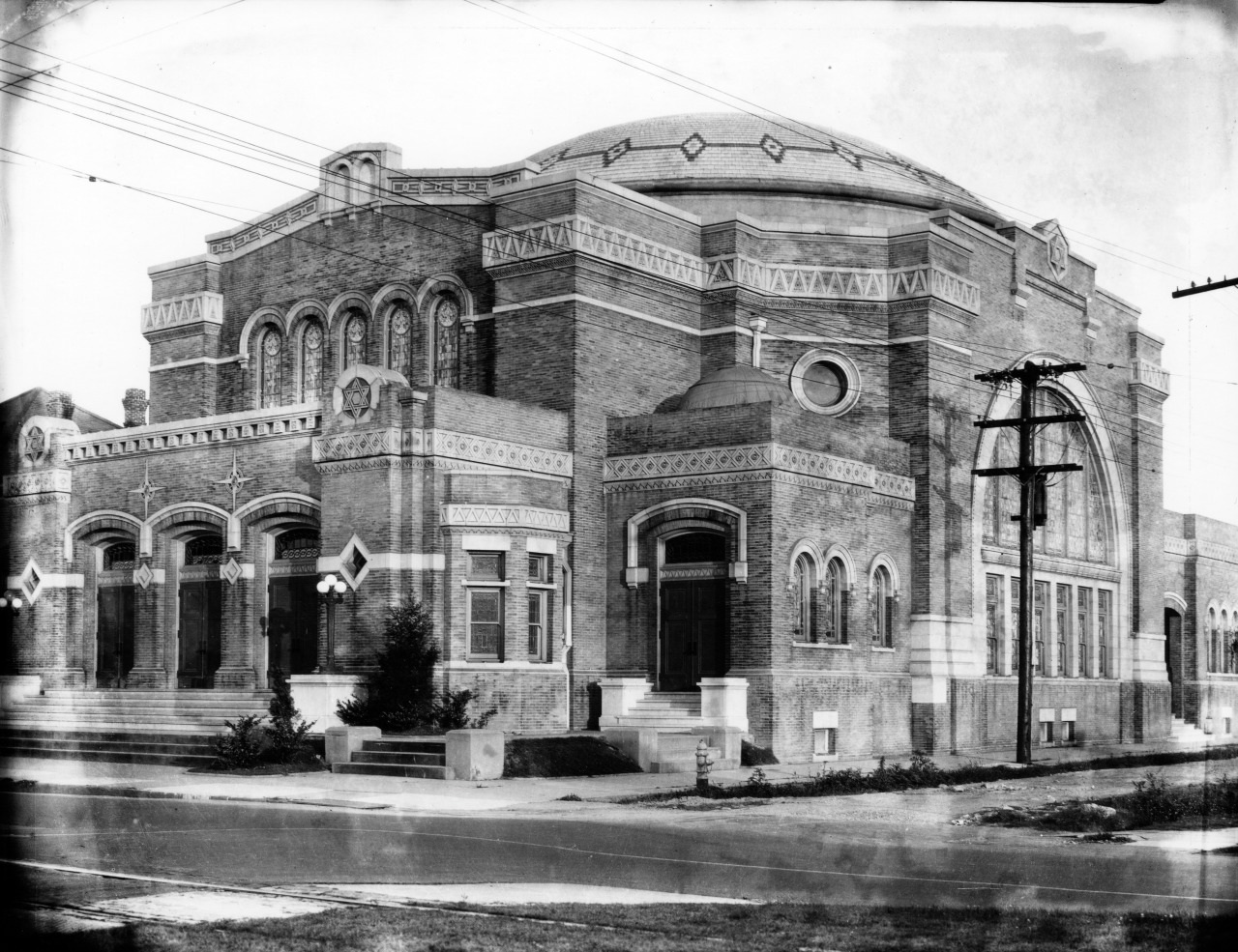 A historic, ornate brick building with arched windows and intricate details, featuring a large dome on top. The structure has steps leading to its entrance and is situated next to a street lined with utility poles.