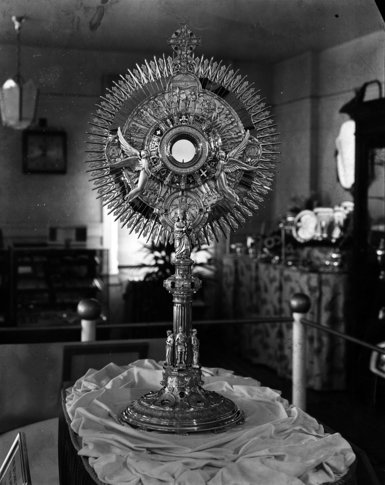 A detailed, ornate religious monstrance stands on display, featuring intricate metalwork with angels and a central circular opening. It is placed on a cloth-covered surface in a dimly lit room.
