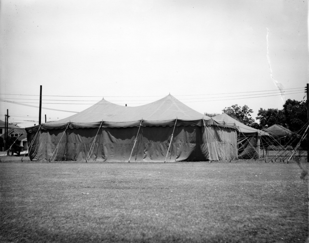 A large canvas tent is set up on a grassy field under a clear sky. The tent is peaked and secured with ropes and poles. Some trees and utility poles are visible in the background.