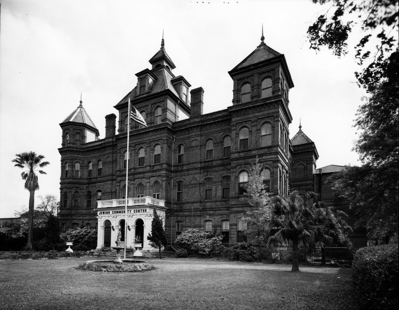 A historic, large brick building with ornate architecture stands prominently. It features multiple towers and a central entrance. In the foreground, theres a flagpole and palm trees, surrounded by well-maintained landscaping.