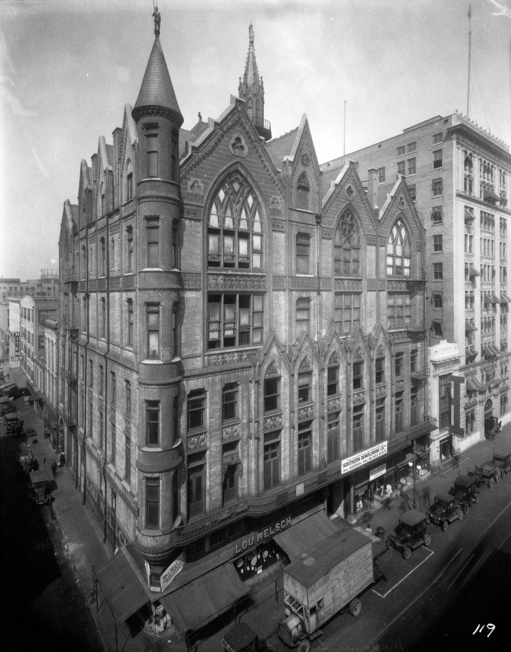 A historic, gothic-style multi-story building on a city street, featuring pointed arches and intricate façade details. Vintage cars and horse-drawn carriages line the street. Large commercial signs are visible at the ground level.