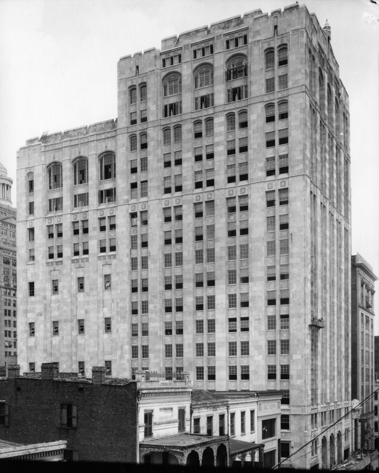 A historic, multi-story building stands tall in an urban setting, featuring an ornate facade with arched windows and intricate architectural details. Other smaller buildings are visible in the foreground, capturing a cityscape from the early 20th century.