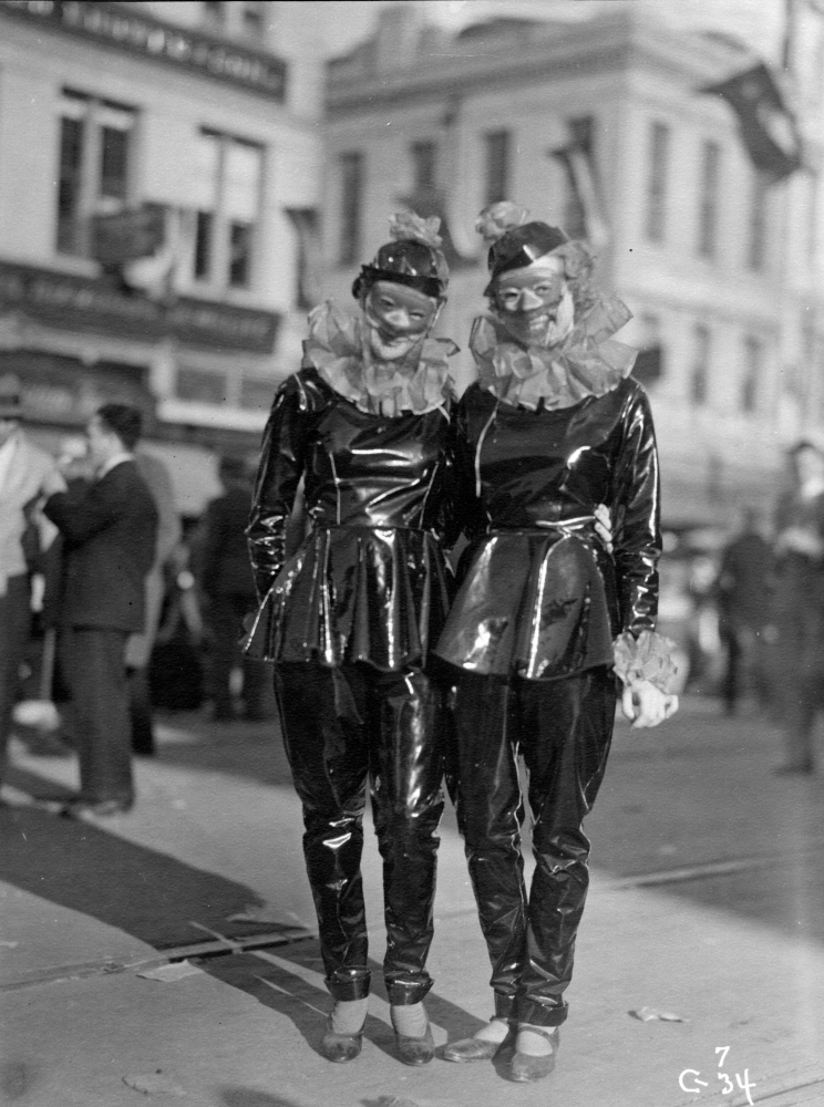 Two people in vintage clown costumes stand side by side on a city street. They wear shiny outfits with ruffled collars and matching hats. The background shows buildings and a crowd of people in early 20th-century attire.