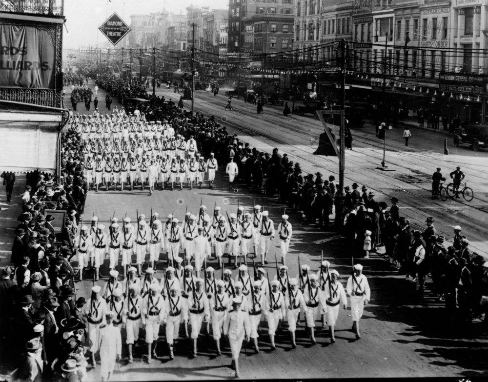 A black-and-white photo of a large parade on a city street. Participants in uniform march in rows, surrounded by spectators on the sidewalk. Buildings and streetcar tracks are visible in the background.