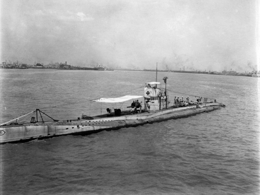 A vintage photograph of a submarine sailing on a calm sea. The submarine surfaces near an industrial shoreline, with buildings visible in the hazy background. Crew members are seen standing on the deck under a canopy.