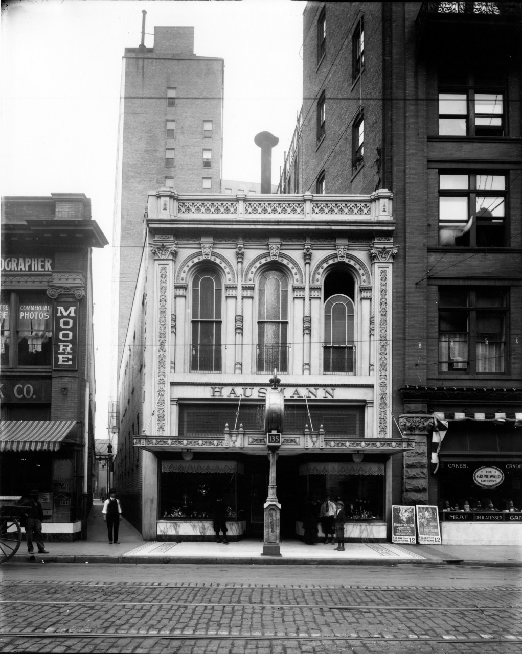 Black and white vintage photo of Hausmann storefront, featuring an ornate two-story facade with arched windows, sandwiched between taller city buildings. People stand at the entrance, and signs hang above neighboring shops. Cobblestone street in front.