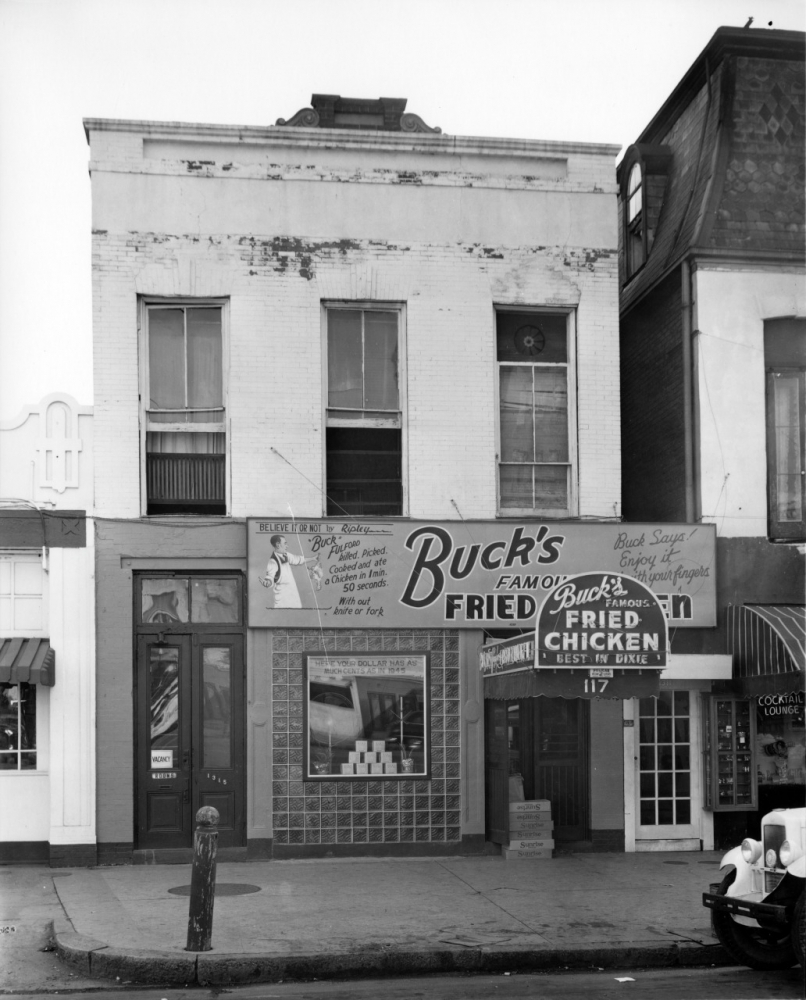 Black-and-white photo of a storefront with a sign reading Bucks Famous Fried Chicken. The building has two floors and a vintage exterior. A sidewalk and fire hydrant are visible in front of the shop.