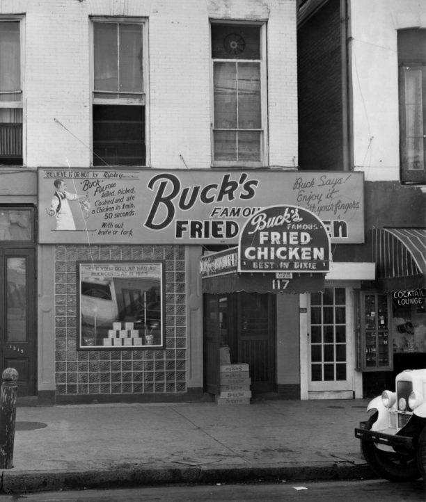 Black and white photo of a storefront with signs for Bucks Famous Fried Chicken, promoting delivery and quality. The building has brick walls, tiled windows, and a vintage car is parked nearby.
