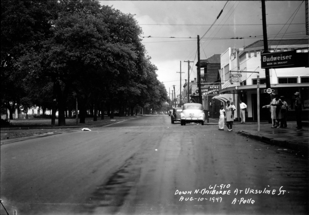 Black and white photo of a street view in 1947. A few people walk along the sidewalk near a bar with a Budweiser sign. A vintage car is parked on the road. Large trees line the street. Handwritten notes indicate location and date in the bottom right.