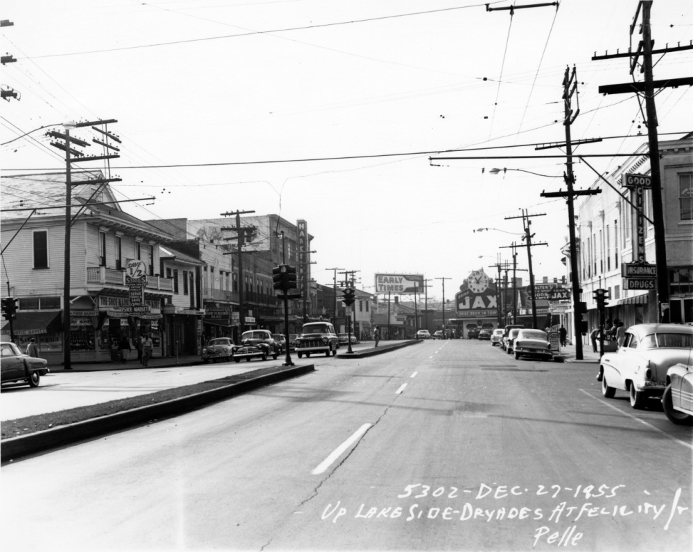 A black-and-white photo of a street scene from December 1955, featuring cars parked along the road and various storefronts. Signs for JAX Beer and an Early Bird Cafe are visible. Power lines and utility poles line the street.