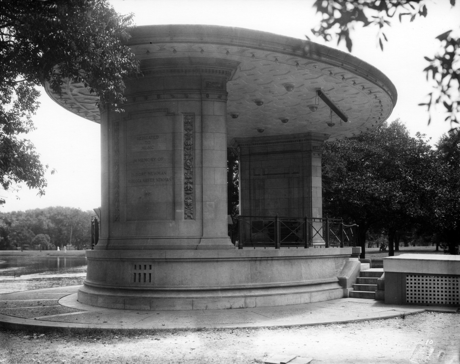 Black and white photo of a large, circular stone bandstand with ornate details. The structure is surrounded by trees and overlooks a body of water. The scene is peaceful, with no people present.